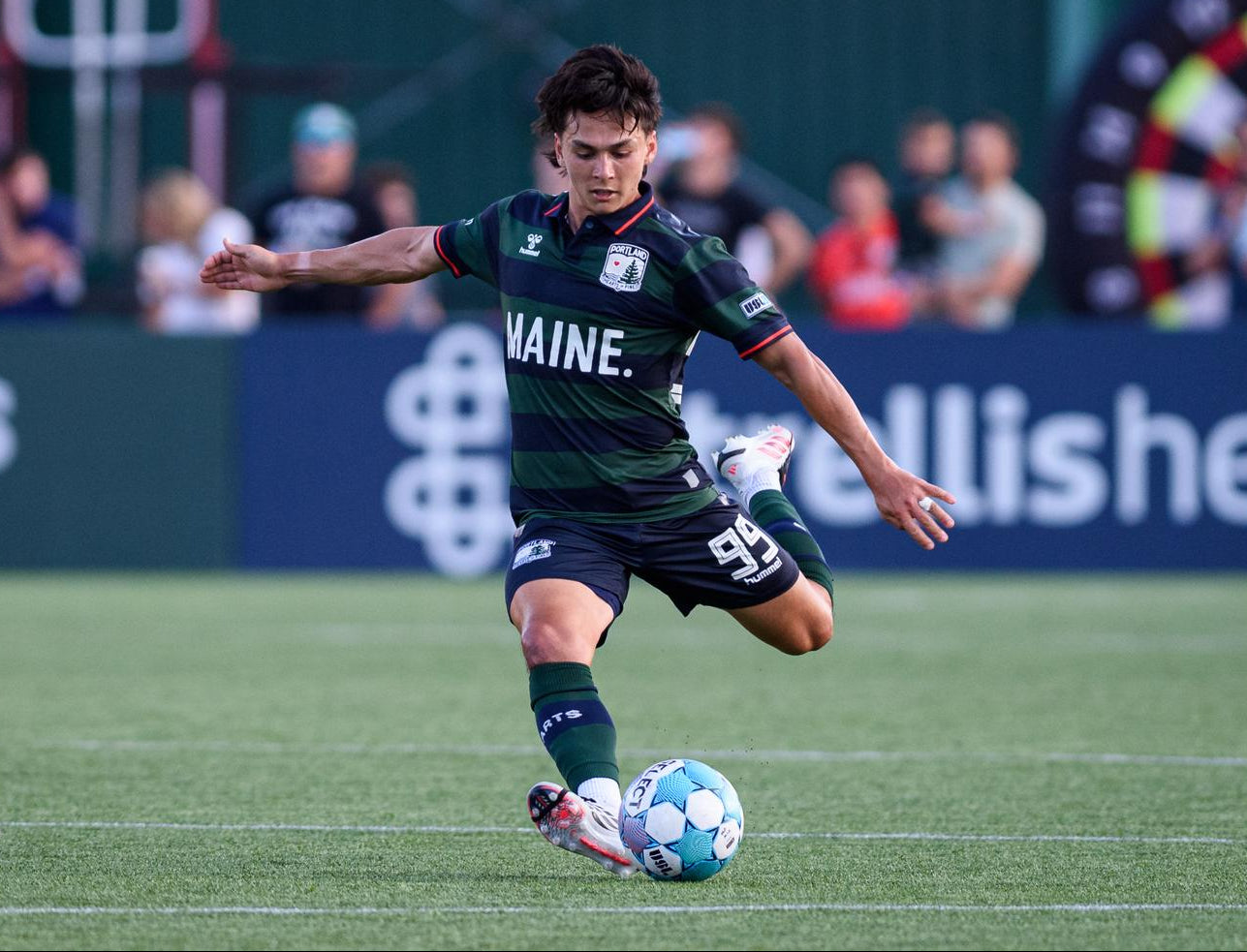 A soccer player in a dark green and black Maine jersey kicks a blue and white soccer ball on a green field, with another player in a light blue jersey partially visible on the left and stadium advertising boards in the background.