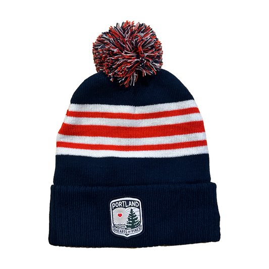 A striped beanie hat with a pom-pom, featuring navy blue and red stripes with a white section that includes a woven Hearts Crest patch.