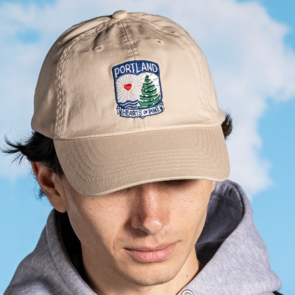 A person wearing a beige 'Hearts of Pine Dad Hat' with navy detailing and an embroidered patch on the front that reads 'PORTLAND Hearts of Pine'.