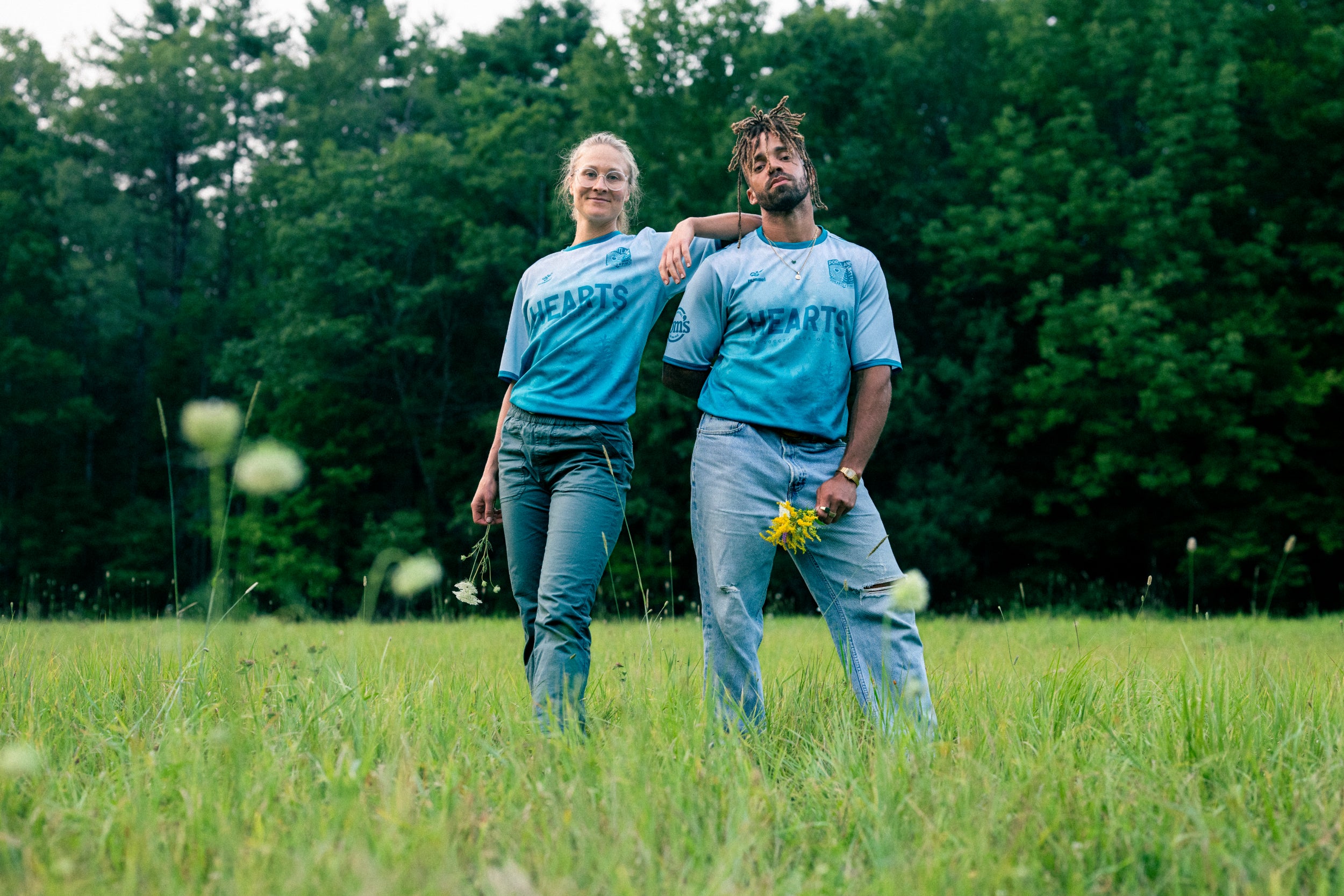 Two people in matching blue shirts standing in a grassy field with trees in the background.